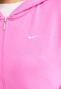Felpa con zip rosa realizzata in tessuto morbido, con un piccolo logo bianco della Nike sul lato sinistro del petto. Include un cappuccio e zip frontale.