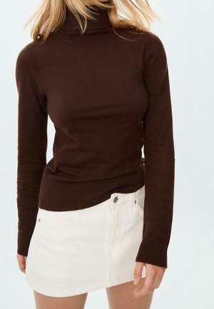 Vrouw die een bruin langarmig coltrui draagt en een witte mini-rok met knopen, staand tegen een effen lichte achtergrond.