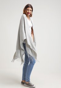Light gray poncho with fringed edges, made from soft fabric. Worn over a white top and paired with blue jeans and gray sneakers.