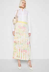 Weißer, plissierter Maxirock mit floralem Druck in Pink und Gelb, kombiniert mit einem weißen Hemd und einer beigefarbenen Handtasche. Model trägt weiße High Heels.