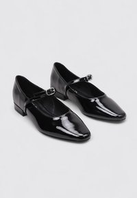 Zapatos de charol negro con puntera cuadrada y tacón bajo grueso, con una fina correa y un detalle de hebilla rectangular.