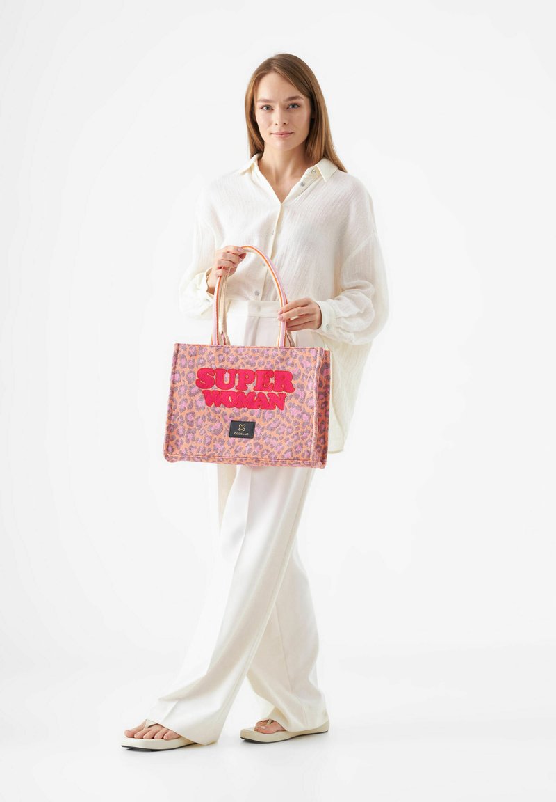 Roze luipaardprint totebag met oversized "SUPER WOMAN" lettering, dubbele handvatten en een zwart merklabel. Gladde textuur, gestructureerde vorm.