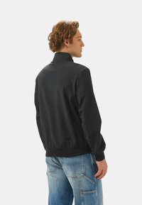 Giubbotto bomber nero con colletto rialzato, polsini e orlo a coste e una texture liscia. Abbinato a jeans in denim chiaro.