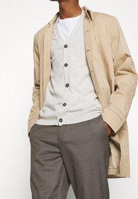 Beige-Mantel, graue Strickjacke mit sechs Knöpfen, weißes Unterhemd und karierten braunen Hosen. Der Stoff wirkt leicht und strukturiert.