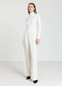 Weißes Rollkragenoberteil kombiniert mit hoch taillierten, weiten cremefarbenen Hosen. Beide aus glattem Stoff, ergänzt durch schwarze spitze Pumps.