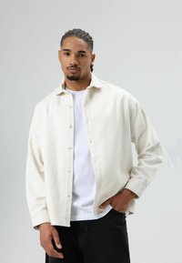 Wit overhemd met knopen, een zachte textuur, voorzien van een kraag, knopen aan de voorkant en lange mouwen, gedragen over een wit T-shirt en donkere spijkerbroek.