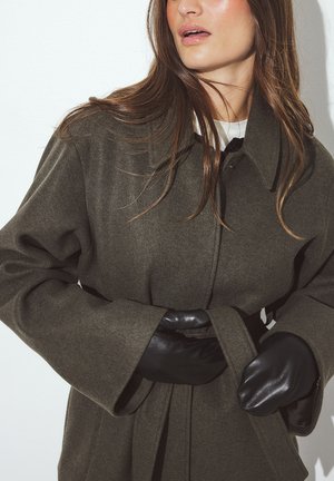 Olive zaļa vilnas mētelis ar apkakli, jostu viduklī un platām piedurknēm, kopā ar melnām ādas cimdiem. Gluda tekstūra ar minimālām detaļām.