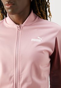 Rožinė užtrauktuku užsegama striukė su standiu apykakle, puikiai matoma balta Puma logotipu. Pagaminta iš lygaus audinio, kontrastingų tamsių rankovių.