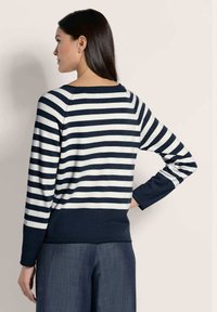 Frau mit langen dunklen Haaren trägt ein dunkelblau-weiß gestreiftes Langarmoberteil und dunkelblaue weite Hosen, mit dem Rücken zur Kamera.