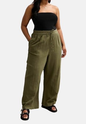 Vrouw draagt een zwarte top zonder bandjes, losse olijfgroene broek met trekkoord en zwarte sandalen met gespen, staand met één hand in de zak.