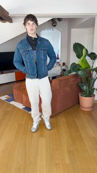 Traper jakna s čavlima preko crne majice, bijelih trenirki i srebrnih tenisica. Unutarnji prostor s biljkom i retro sofom.