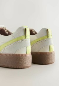 White and cream sneakers with a textured tan sole, featuring green accents and braided stitching on the rear. Soft materials used throughout.