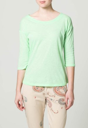 Lichtgroen, losvallend shirt met lange mouwen en een wijde halslijn, gecombineerd met beige, patroonbroek met paisley-ontwerpen.