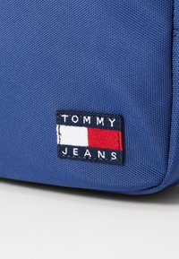 Borsa in tessuto blu con una toppa rettangolare cucita con il testo "TOMMY JEANS" e un design a strisce orizzontali rosso, bianco e blu.