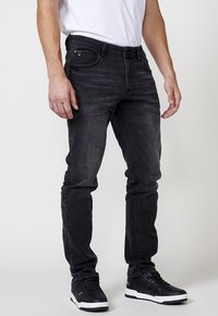 Jeans de mezclilla negra ajustada con cinco bolsillos, sutil desgaste y costuras tonales. Combinados con zapatillas negras y una camisa blanca.