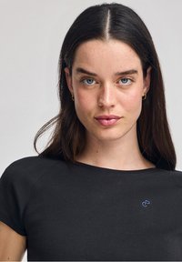 Svart t-shirt med kort ärm och rund halsringning, med en liten blå logotyp på bröstet. Slät textur och figurnära design.