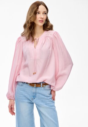 Kobieta o falowanych włosach nosi jasnoróżową bluzkę z długim rękawem z haftowanymi ramionami oraz jasnoniebieskie dżinsy, stojąc na tle białego tła.