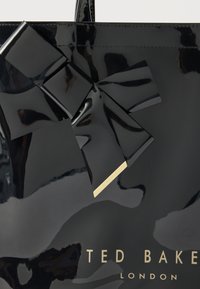 Γυαλιστερή μαύρη τσάντα Ted Baker London με μεγάλο λαμπερό φιόγκο που διαθέτει μια μικρή χρυσή ταυτότητα.