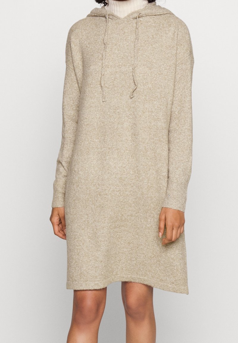 Femme portant une robe-pull à capuche beige en tricot à manches longues, sur un col roulé blanc, se tenant devant un fond clair uni.