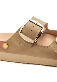 Sándalo beige con un acabado suave de ante, que presenta dos correas ajustables y una gran hebilla metálica. Suela texturizada con pequeñas protuberancias.