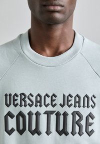 Světle šedá mikina s vyraženým černým textem "VERSACE JEANS COUTURE", s ribbovaným véčkovým límcem a raglánovými rukávy.