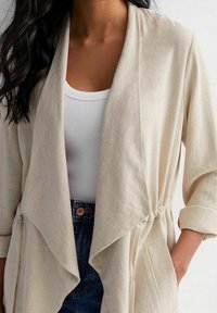 Cárdigan beige ligero con frente drapeado, mangas 3/4 y lados fruncidos; usado sobre una camiseta sin mangas blanca de canalé y vaqueros.