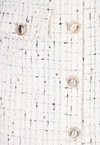 Hvid tekstureret stof med sorte og gyldne tråde, der har en lille brystlomme og runde sølvknapper langs fronten.