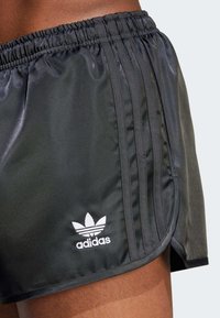 Schwarze Sportshorts mit glänzender Oberfläche, ausgestattet mit einem elastischen Bund, drei vertikalen Streifen und einem weißen Adidas-Logo am Saum.