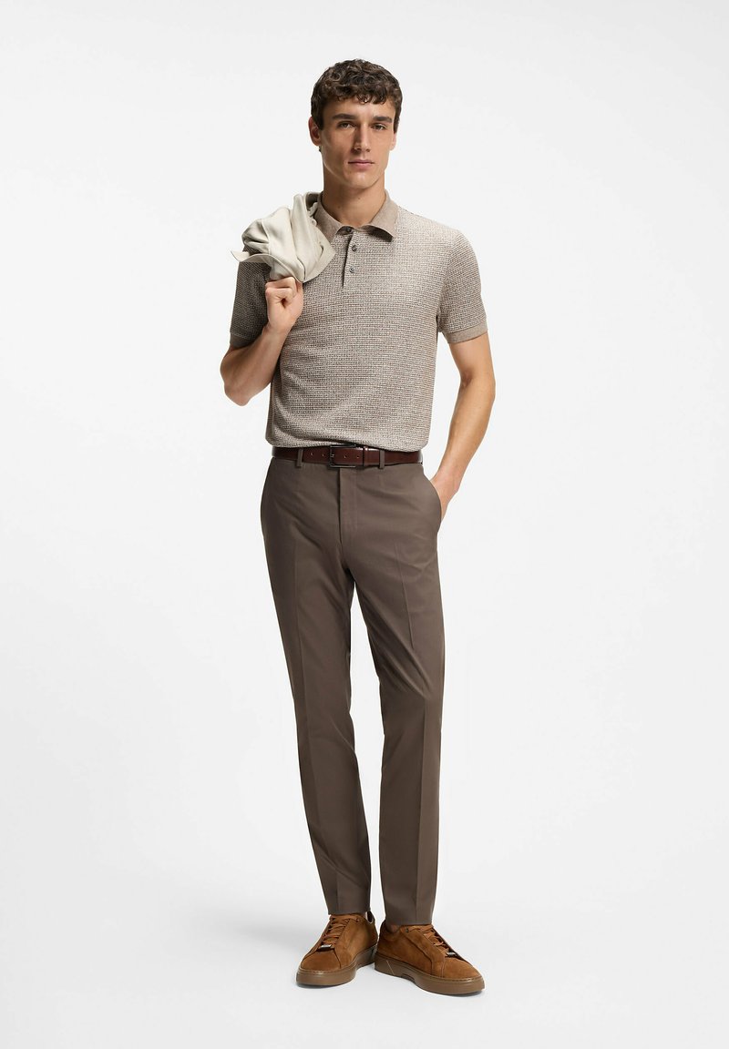Homme portant un polo en maille beige, un pantalon marron et des baskets marron, tenant une veste légère sur son épaule sur un fond blanc.