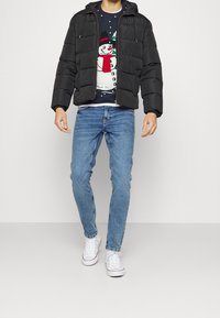 Zwarte pufferjack met een capuchon, rits aan de voorkant en gestikte details, gedragen over een marineblauw sneeuwpoptrui, gecombineerd met blauwe spijkerbroeken en witte sneakers.