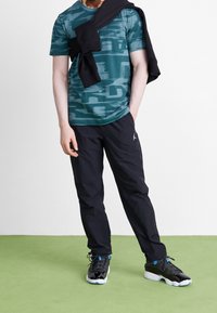 Mann mit teal-farbenem gemustertem T-Shirt, schwarzen Hosen mit Logo, Sneakern und schwarzem Hoodie, der über den Schultern drapiert ist, steht auf grünem Boden.