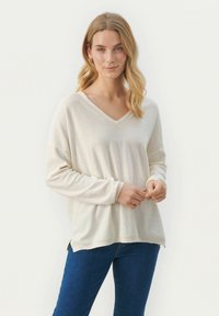 Cremfarbener Pullover mit V-Ausschnitt und langen Ärmeln, weichem Material sowie seitlichen Schlitzen, kombiniert mit blauen Jeans. Der Stoff wirkt leicht und lässig.