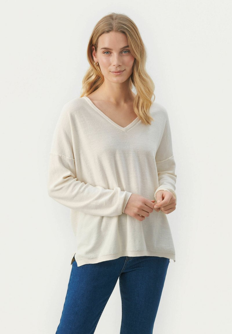 Cremfarbener Pullover mit V-Ausschnitt und langen Ärmeln, weichem Material sowie seitlichen Schlitzen, kombiniert mit blauen Jeans. Der Stoff wirkt leicht und lässig.