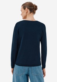 Tummansininen pitkähihainen neule, jossa on pyöreä pääntie, valmistettu pehmeästä kankaasta. Varustettu joustinvärillä helmassa ja hihansuissa, yhdistettynä vaaleansinisiin farkkuihin.