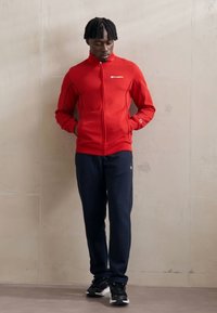 Chaqueta roja de cremallera con acentos negros, combinada con pantalones de chándal azul marino. Zapatillas negras completan el conjunto atlético. Textura de tela suave.