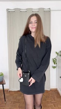 Black long-sleeve dress, with a loose fit. Holding a small black crossbody bag featuring a heart-shaped accent. Room with minimal decor.