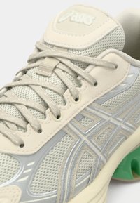 Light gray athletic shoe with mesh and synthetic upper, reflective silver accents, beige laces, and a green sole. Features a textured design.