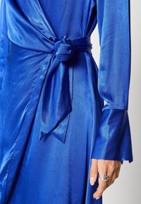 Světle modré saténové šaty s opaskem na uzel a rozšířenými rukávy, na fotografii je ruka, která nosí moderní stříbrný prsten.