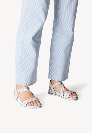 Sandales gris clair avec plusieurs brides croisées, semelle plate et détails de couture subtils, associées à un pantalon rayé bleu et blanc.