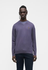 Maglione viola a scollo a V con lavorazione a maglia testurizzata, caratterizzato da un piccolo logo giallo sul lato sinistro del petto. Indossato con pantaloni scuri, design minimalista.