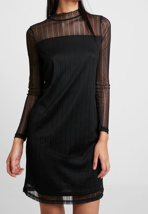 Žena v černých, žebrovaných, dlouhých rukávech, s průhlednou proužkovanou látkou na rukávech, výstřihu a lemu, stojící před jednobarevným pozadím.