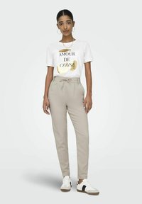 T-shirt blanc avec un texte graphique doré, associé à un pantalon beige clair sur mesure avec taille à cordon et des baskets blanches avec des accents noirs.