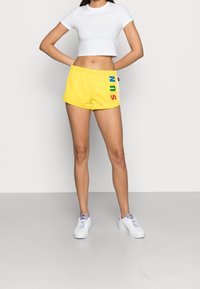 Žluté sportovní šortky s nápisem "SUN" v duhových barvách po boku, doplněné bílým přiléhavým tričkem a bílými teniskami.