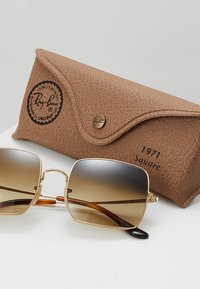 Ochelari de soare Ray-Ban cu lentile degrade maro, cadru din metal auriu și accente de caramel, prezentati cu o carcasa texturata din piele maro.