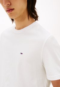 Nahaufnahme einer Person, die ein schlichtes weißes T-Shirt mit einem kleinen roten, weißen und blauen rechteckigen Logo auf der Brust trägt.