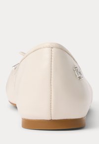 Plat crèmekleurig leren schoen met een bruine zool, zichtbare stiksels aan de achterkant en een klein zilveren decoratief embleem op de hak.