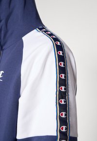 Φούτερ με κουκούλα σε ναυτικό μπλε και άσπρες επιφάνειες, με ριγέ πλευρικό σχέδιο με λογότυπα και μπλε, κίτρινες και μαύρες λεπτομέρειες.