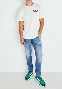 Man står klädd i en vit "LEE"-t-shirt, ljusblå jeans och gröna sneakers mot en helt vit bakgrund.