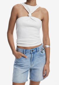Top blanco de tirantes sin mangas con un detalle de nudo en el cuello, combinado con pantalones cortos de mezclilla azul claro que presentan bordados florales en la cinturilla.
