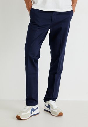 Navy blue trousers with a straight fit, smooth fabric, and two side pockets, paired with white and blue athletic sneakers.